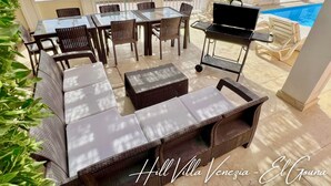 Een terras buiten met rieten meubilair, een barbecue en uitzicht op een zwembad en resortgebouwen.