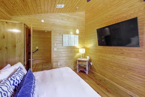 A wooden-paneled room with a bed, a TV mounted on the wall, and a small bedside table with a lamp. - Hot Tub, Fire Pit, Forest Views! Broken Bow Cabin (Broken Bow)