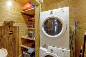 A compact laundry room with a front-loading washing machine and a drying machine, wooden walls, and a shelf with various items. - Hot Tub, Fire Pit, Forest Views! Broken Bow Cabin (Broken Bow)