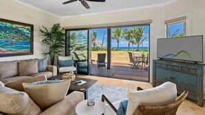A living room with a sofa, chairs, a coffee table, and a TV. There is a large window with a view of the beach and palm trees.