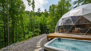 Een hottub met een houten terras en een overdekte zithoek, omgeven door bebost gebied met een grindpad.