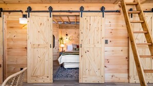 Une cabane en bois dotée de deux grandes portes coulissantes qui donnent sur une chambre à coucher comprenant un lit, une table de chevet et une plante suspendue.