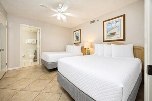 Une chambre d’hôtel avec deux lits, un ventilateur de plafond, une salle de bain avec des toilettes et un lavabo, et des œuvres d’art encadrées accrochées aux murs.