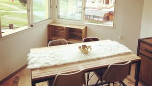 A dining area with a table and chairs, large windows with a view of a restaurant and trees. - South-facing apartment in resort center (Isola 2000)