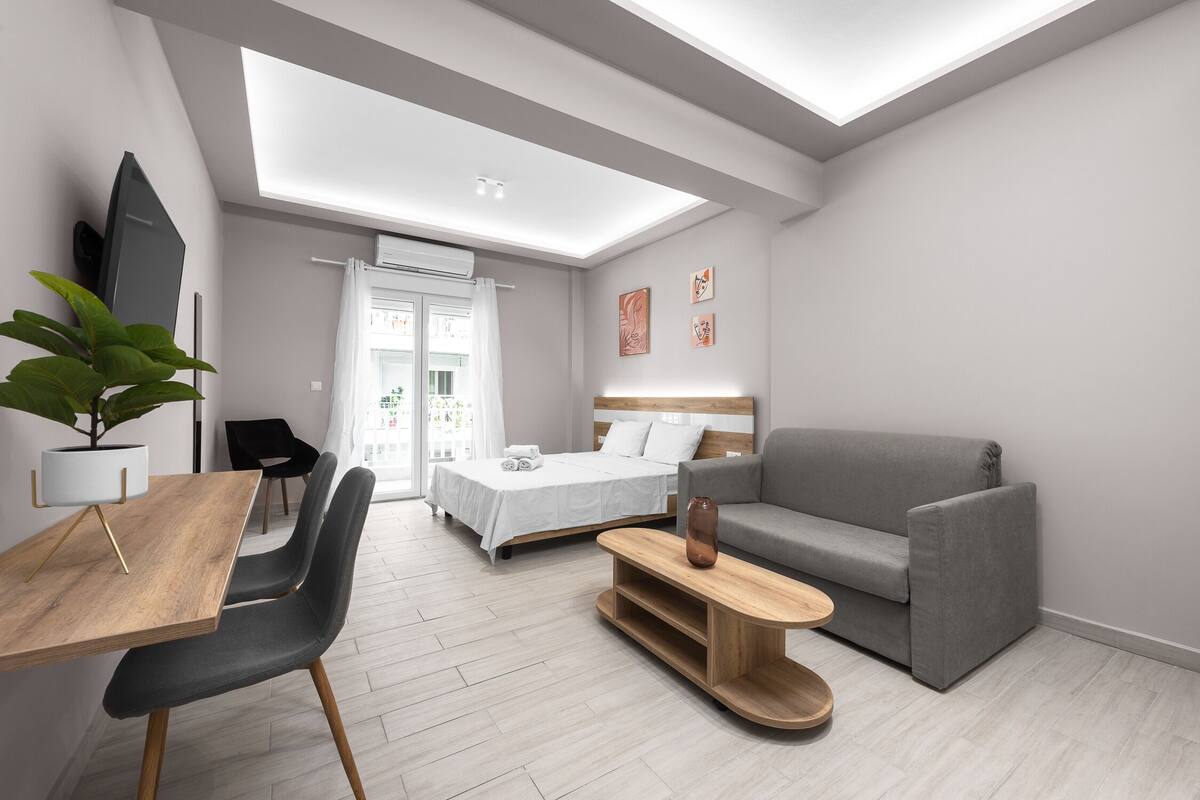 Una habitació moderna i ben il·luminada amb un llit, un sofà gris, una taula de cafè de fusta i una zona de menjador amb taula i cadires.