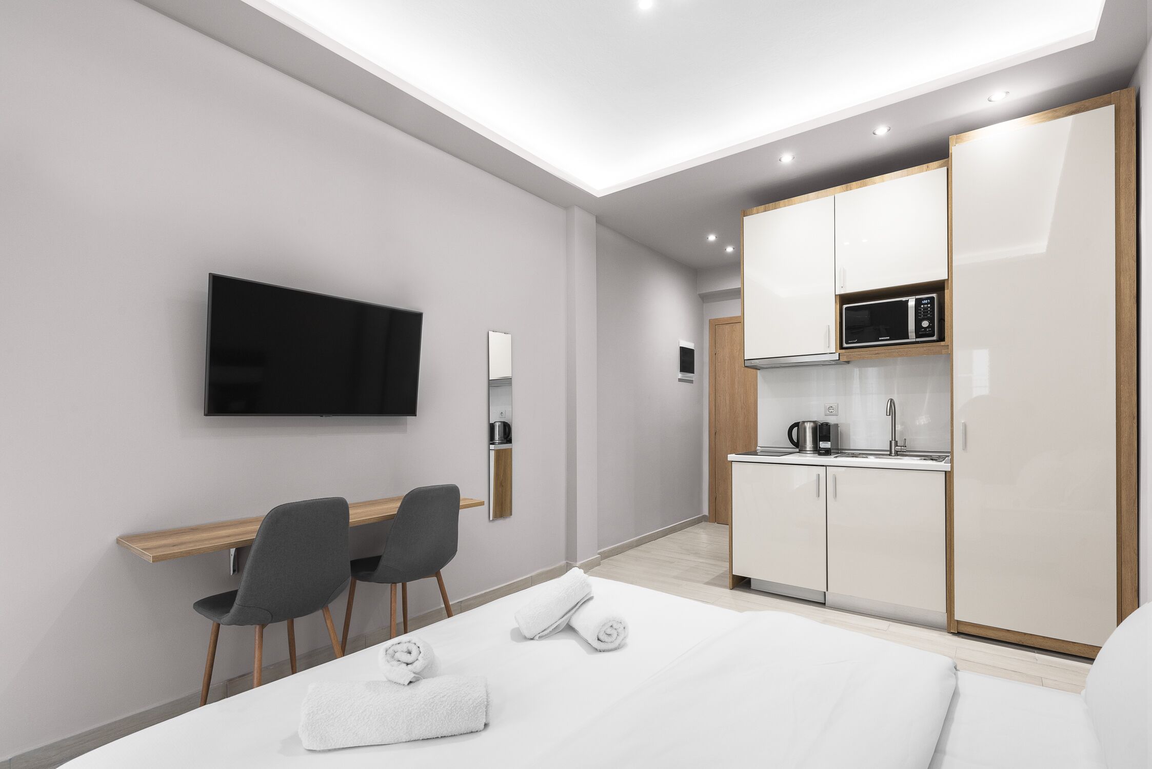 A modern hotel room with a bed, a desk, a chair, a TV, and a kitchenette.