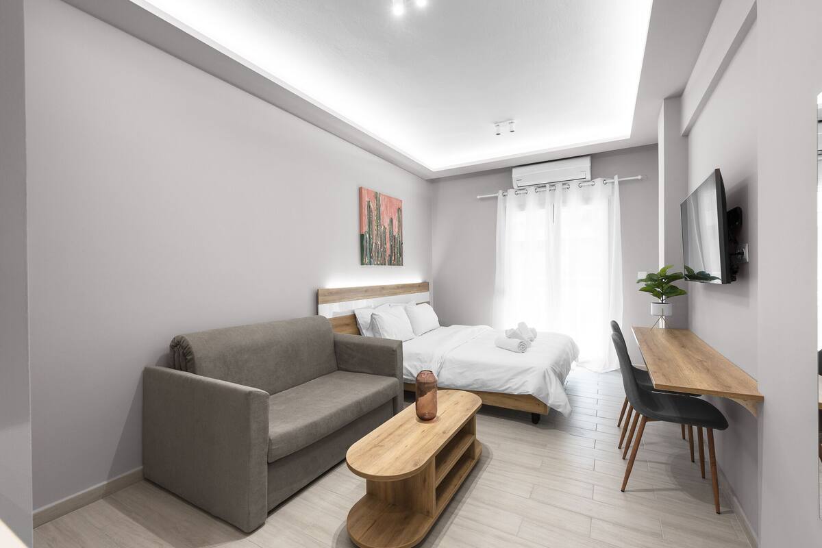 Habitació d'hotel moderna amb un sofà, una taula de centre de fusta, un llit amb roba blanca, un escriptori amb una cadira i un televisor fixat a la paret.