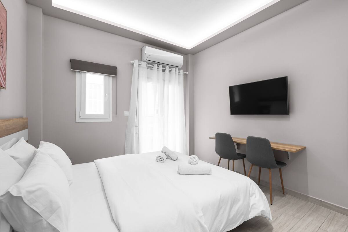 Una habitació d'hotel moderna amb un llit, una taula de treball amb dues cadires, un televisió de pantalla plana i un aparell d'aire condicionat.
