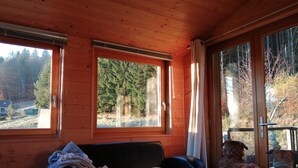 Interior de una cabaña de madera con un sofá de cuero negro, una manta y dos perros mirando hacia el exterior.