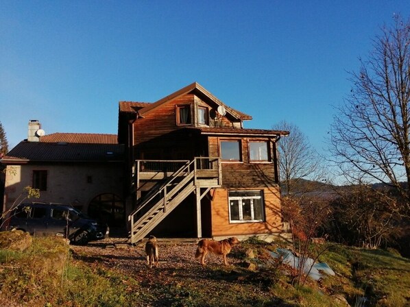 Una casa de madera con una escalera que lleva a un balcón, rodeada de paisaje natural.