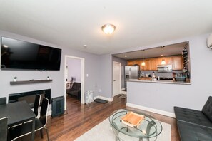 A modern living room with a flat-screen TV, a dining area, a glass coffee table, and a kitchen area with wooden cabinets and a refrigerator.