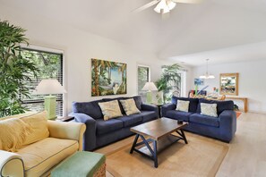 A living room with two sofas, a coffee table, and a small table with a lamp. There are plants, artwork on the walls, and a window with blinds.