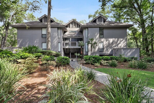 A two-story house with a front porch, surrounded by a well-maintained garden with various plants and a gravel pathway leading to the entrance.