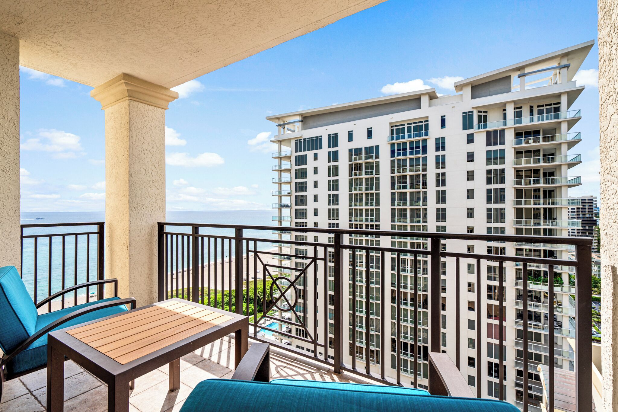 A balcony with a wooden table and blue chairs, overlooking a high-rise building and the ocean.