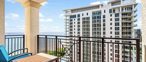 A balcony with a wooden table and blue chairs, overlooking a high-rise building and the ocean.
