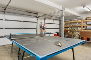 A ping pong table in a garage with a metal rack and various tools and equipment in the background.