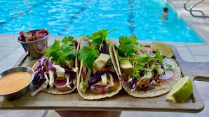 Three tacos on a wooden board with a pool in the background.