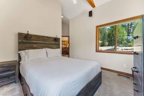 Une chambre avec une tête de lit en bois, une grande fenêtre donnant sur les arbres, un placard intégré et un sol recouvert de moquette.