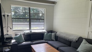 A living room with a dark gray sofa, a wooden coffee table, and a large window with a view of trees. - Waterfront Retreat with Kayaks (Inverness)