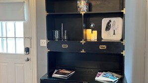 Une pièce avec un bureau noir, un tabouret en bois, une bibliothèque agrémentée d’objets décoratifs et une porte blanche.