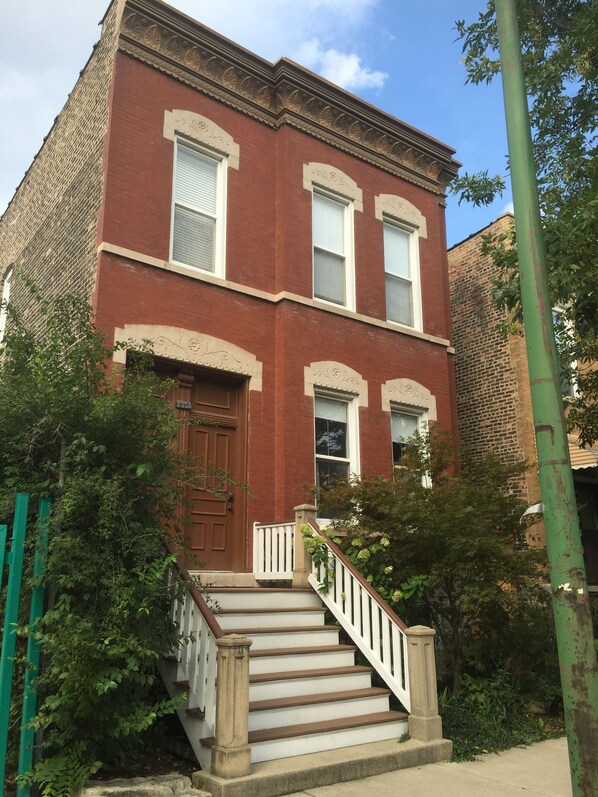 A two-story brick building with a front entrance, a staircase with a white railing, and a small porch area.