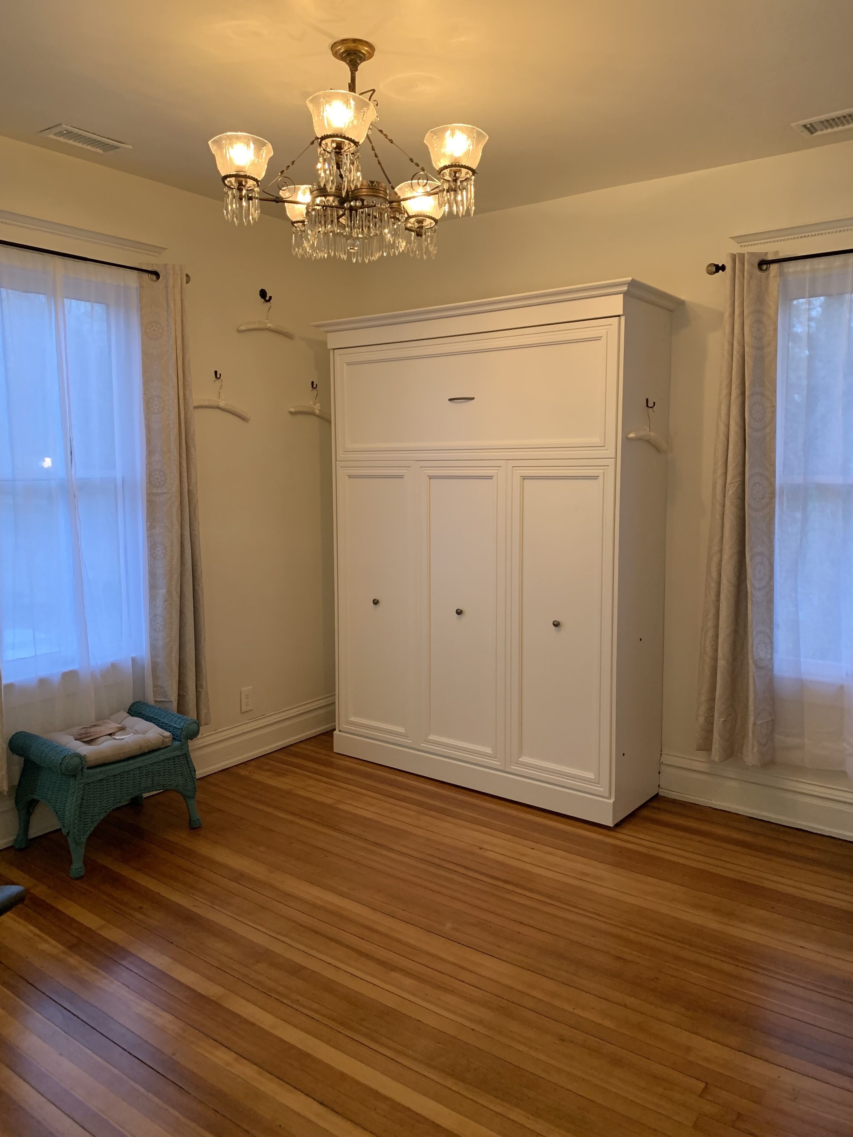 Une pièce avec un plancher en bois, une armoire blanche, un petit banc de couleur turquoise et un lustre.