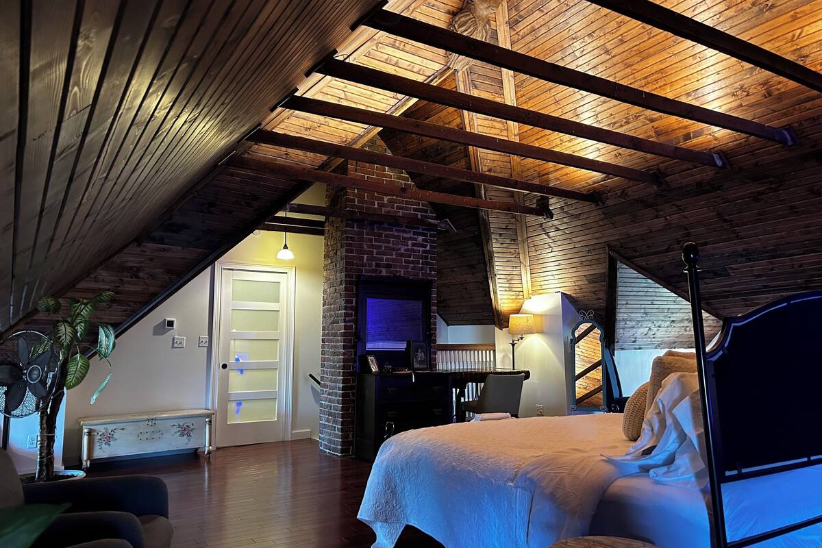 Um quarto de sótão acolhedor, com teto inclinado, lareira de tijolos, cama e escrivaninha.