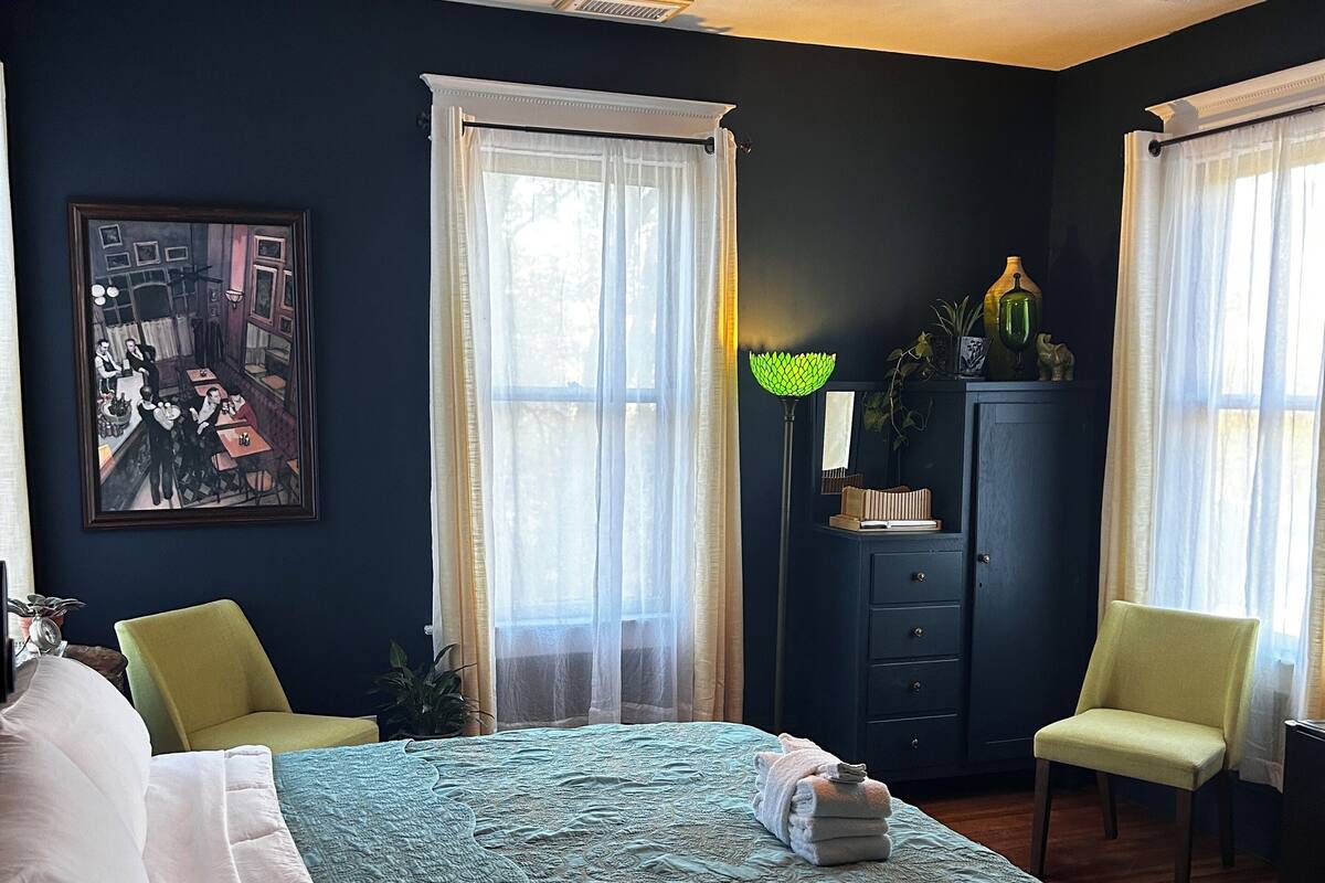 Quarto com cama, duas cadeiras, ventoinha no teto, mesa de cabeceira, abajur, armário e um quadro na parede.