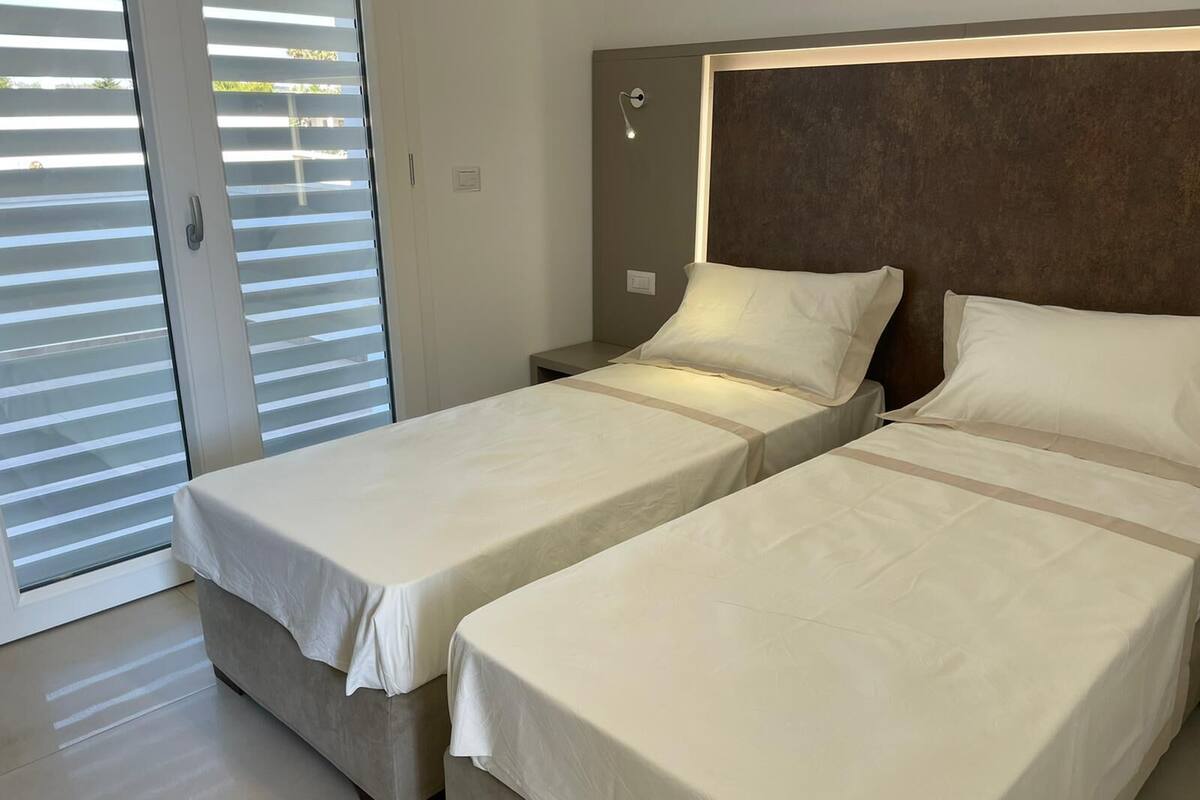 A hotel room with two single beds, a nightstand, a lamp, and a sliding glass door leading to a balcony.