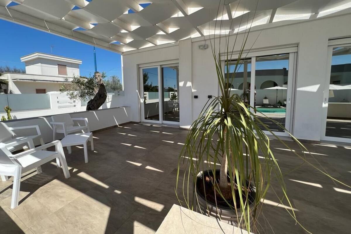 A covered outdoor area with white chairs, a potted plant, and a view of a building in the background.