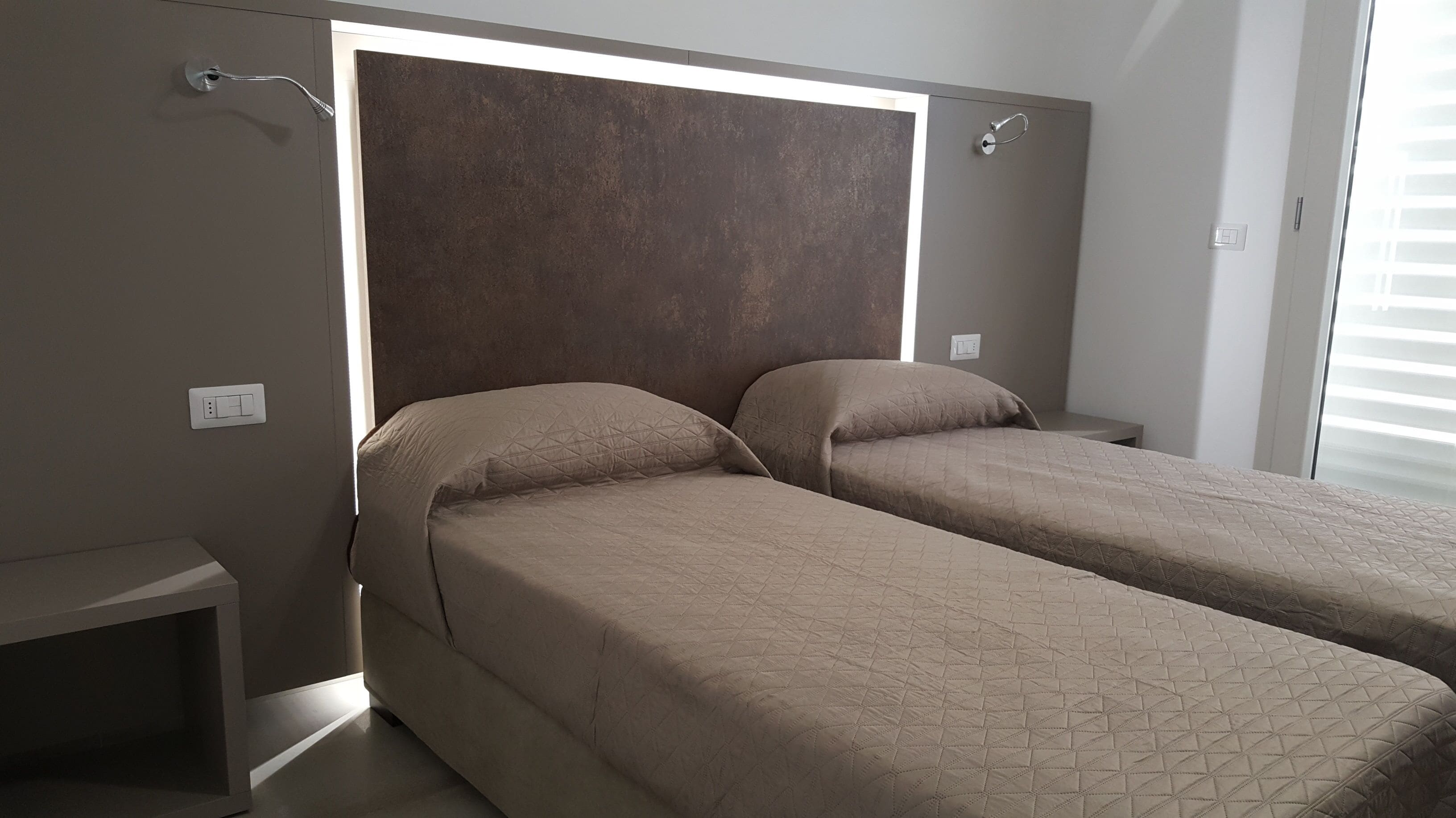 Un dormitorio con dos camas individuales, una mesita de noche, una lámpara de pared y una ventana con persianas.