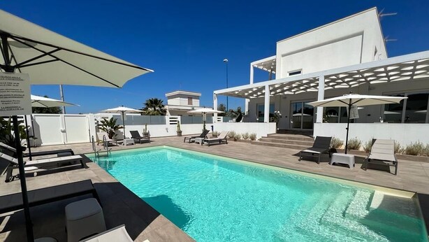 Un área de piscina moderna con sillones de descanso y sombrillas, una piscina azul cristalina y un edificio contemporáneo con fachada blanca.