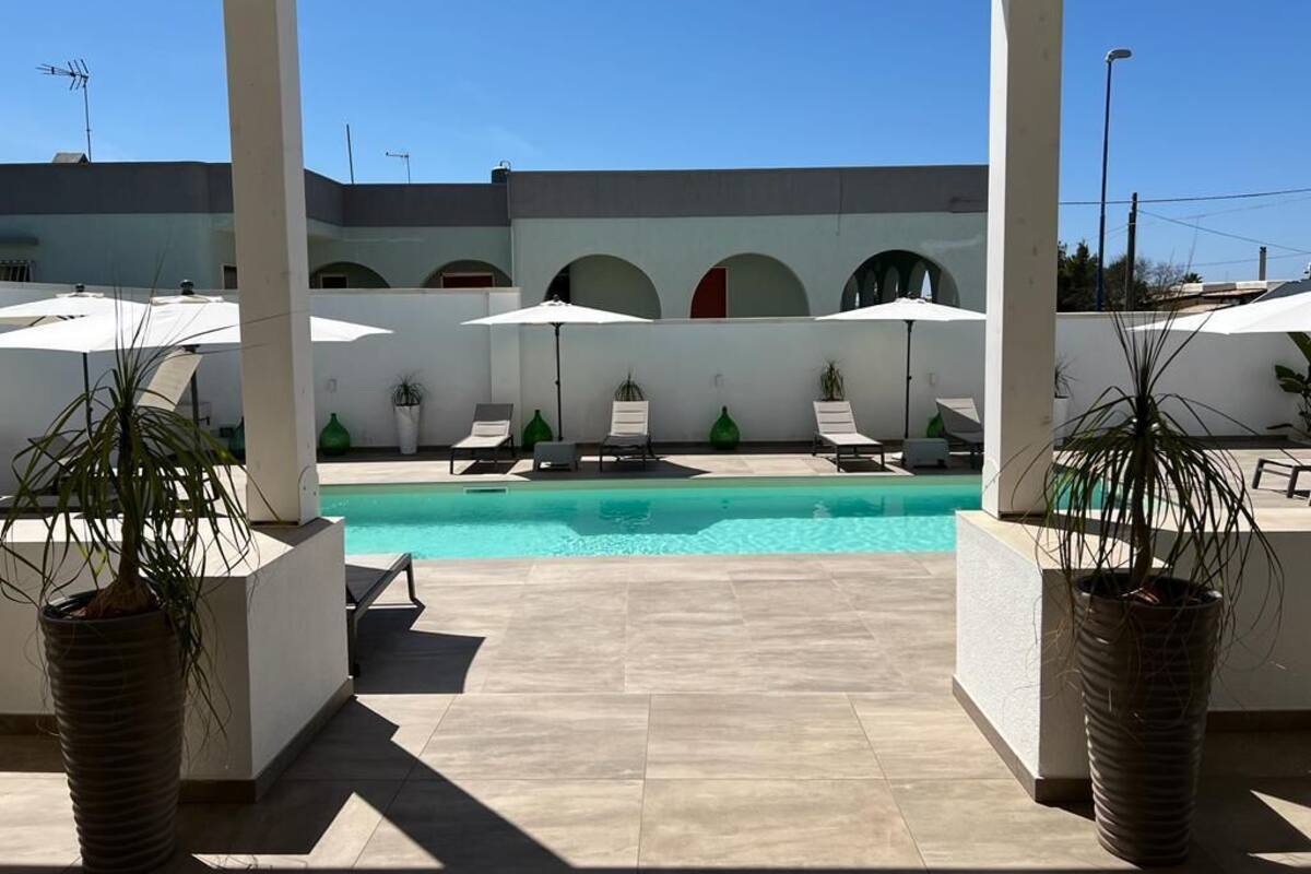 A poolside area with lounge chairs and umbrellas, a clear blue sky, and a building with arches in the background.