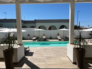 A poolside area with lounge chairs and umbrellas, a clear blue sky, and a building with arches in the background. - Elite Luxury Residence (Racale)