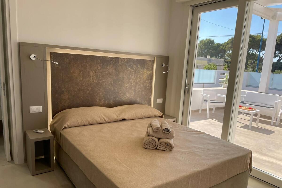 A modern bedroom with a large bed, a small bedside table, and a view of an outdoor patio with seating.