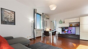 A modern living room with a grey sofa, a dining area with a table and chairs, and a kitchen area with cabinets and a television. - Bright apartment in Via Ginori, in the heart of the Testaccio district (Rome)
