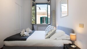 A bedroom with a bed, a nightstand, a lamp, a window with a view of buildings, and a door. - Bright apartment in Via Ginori, in the heart of the Testaccio district (Rome)