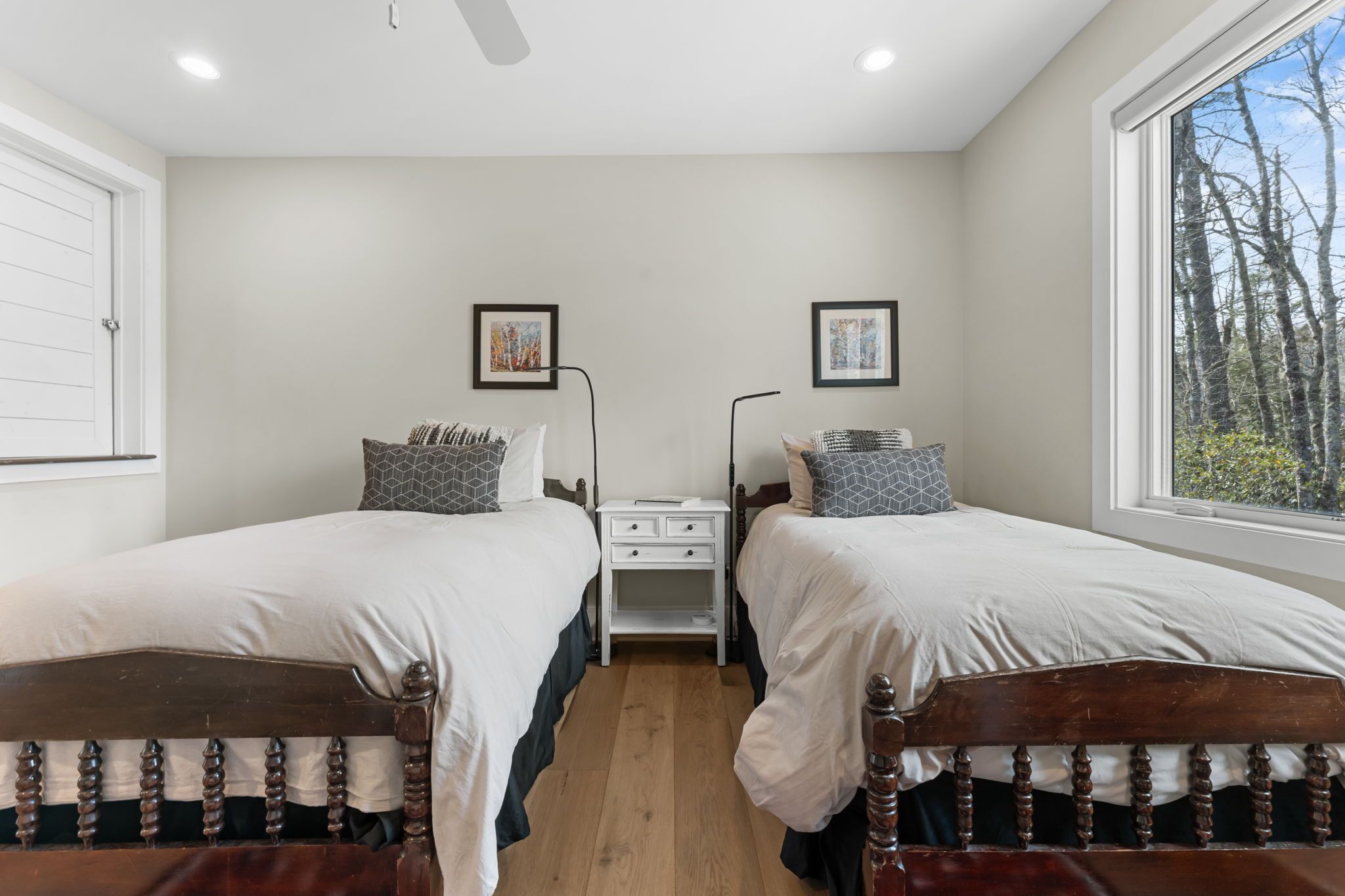 A room with two beds, a nightstand, and a window with a view of trees.
