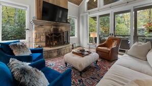 A spacious living room with a stone fireplace, large windows, and a high ceiling.
