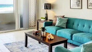 A living room with a teal sofa, a coffee table, and a lamp. There is a view of the beach through a sliding door.