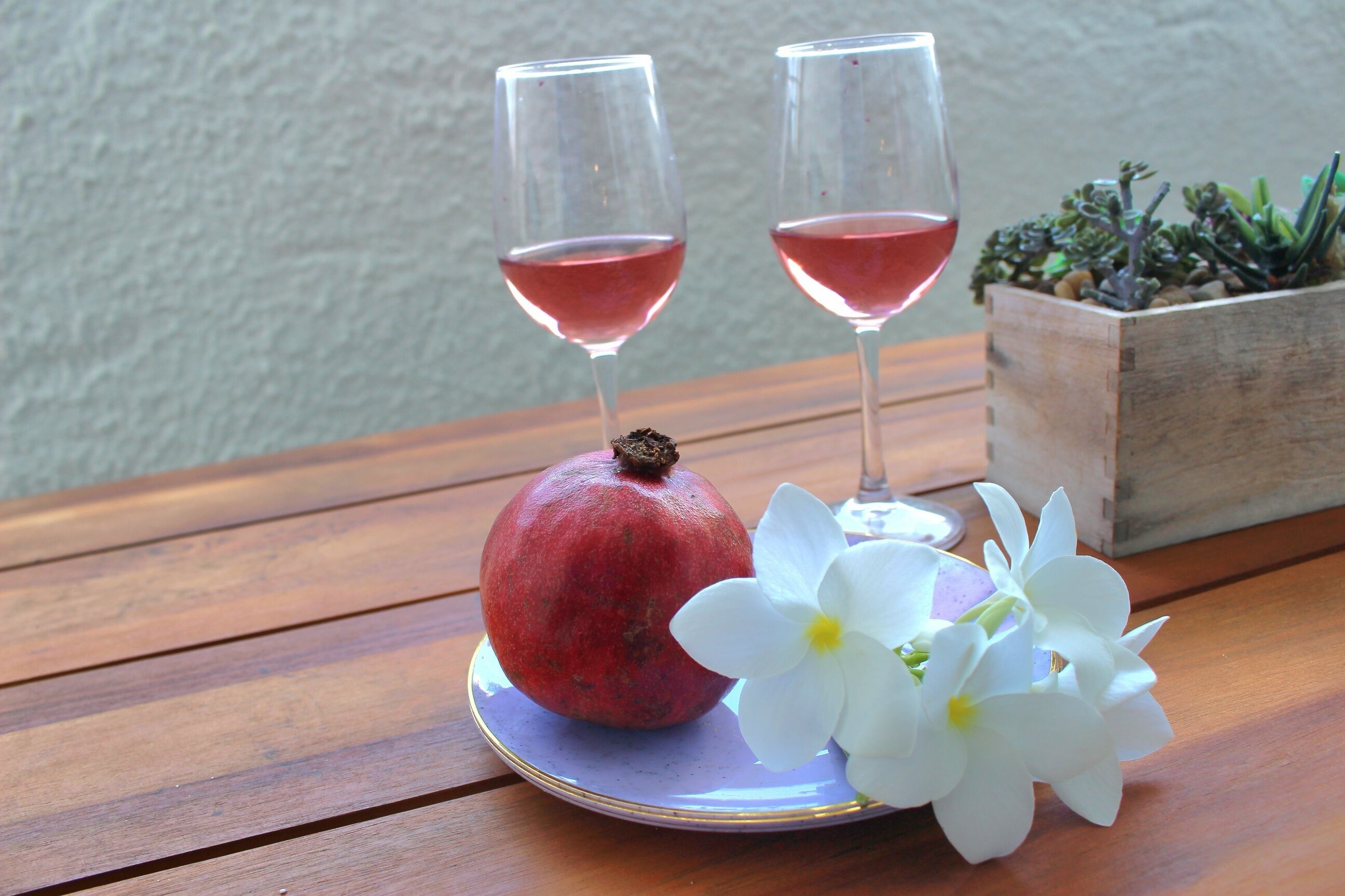 Une table en bois avec deux verres de vin rouge, une grenade et des fleurs blanches.