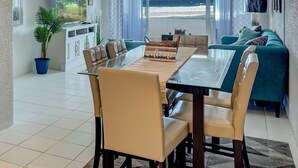 A modern living room with a glass dining table, beige chairs, a teal sofa, and a view of the ocean.