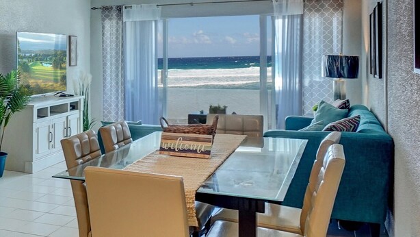 A modern living room with a glass table and chairs, a sofa, and a view of the beach.