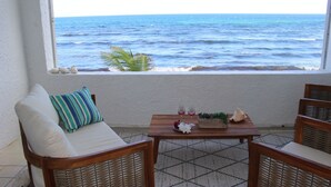 A room with a view of the ocean, a wooden table with a vase and two glasses, and wicker furniture.