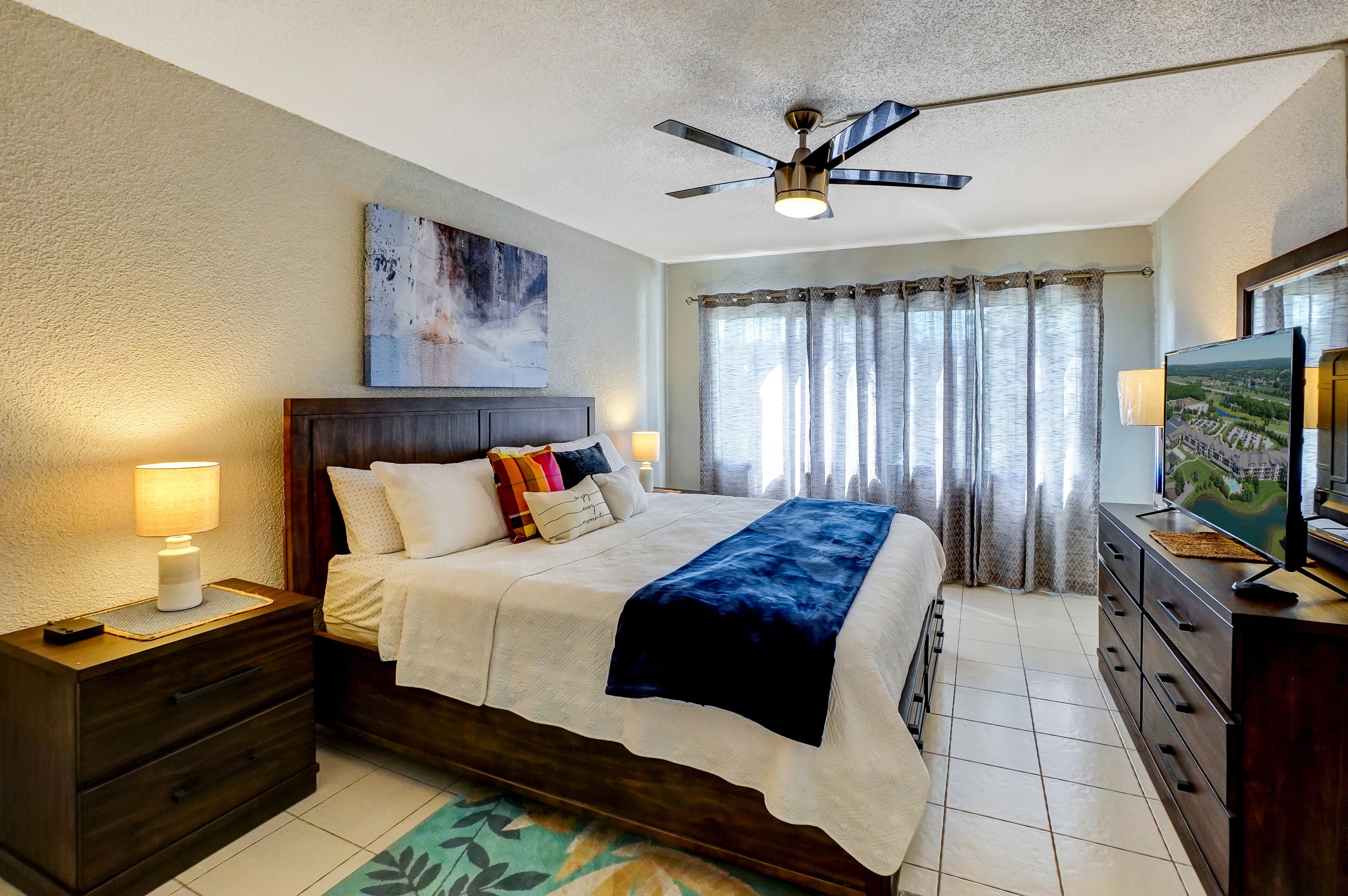 A bedroom with a bed, nightstands, a TV, a ceiling fan, and a window with curtains.