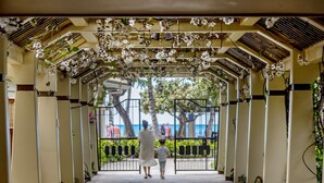 Un paseo techado con suelo de baldosas, bordeado por columnas y plantas, que conduce a una zona vallada con vistas a palmeras y al mar.