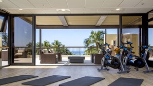 Un gimnasio con bicicletas estáticas y colchonetas, con vistas a un balcón con mobiliario de mimbre y una vista de palmeras.