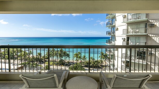 Un balcon avec deux chaises et une petite table, donnant sur une plage bordée de palmiers et sur un océan bleu et limpide.