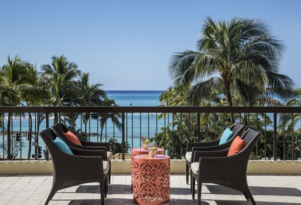 A balcony with wicker chairs and a table, overlooking a beach with palm trees and the ocean.