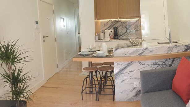 A modern kitchen with marble countertops, a built-in oven, and a coffee machine. There is a bar area with stools and a grey sofa with red and white cushions in the living area. - Principe Real Central Home - Free Airport Transfer (Lisboa)